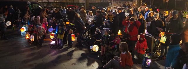 Ev.-luth. Epiphaniengemeinde Hamburg – Laternenumzug Große Gruppe von Kindern und Erwachsenen in der Dunkelheit mit leuchtenden Laternen auf dem Kirchenvorplatz.